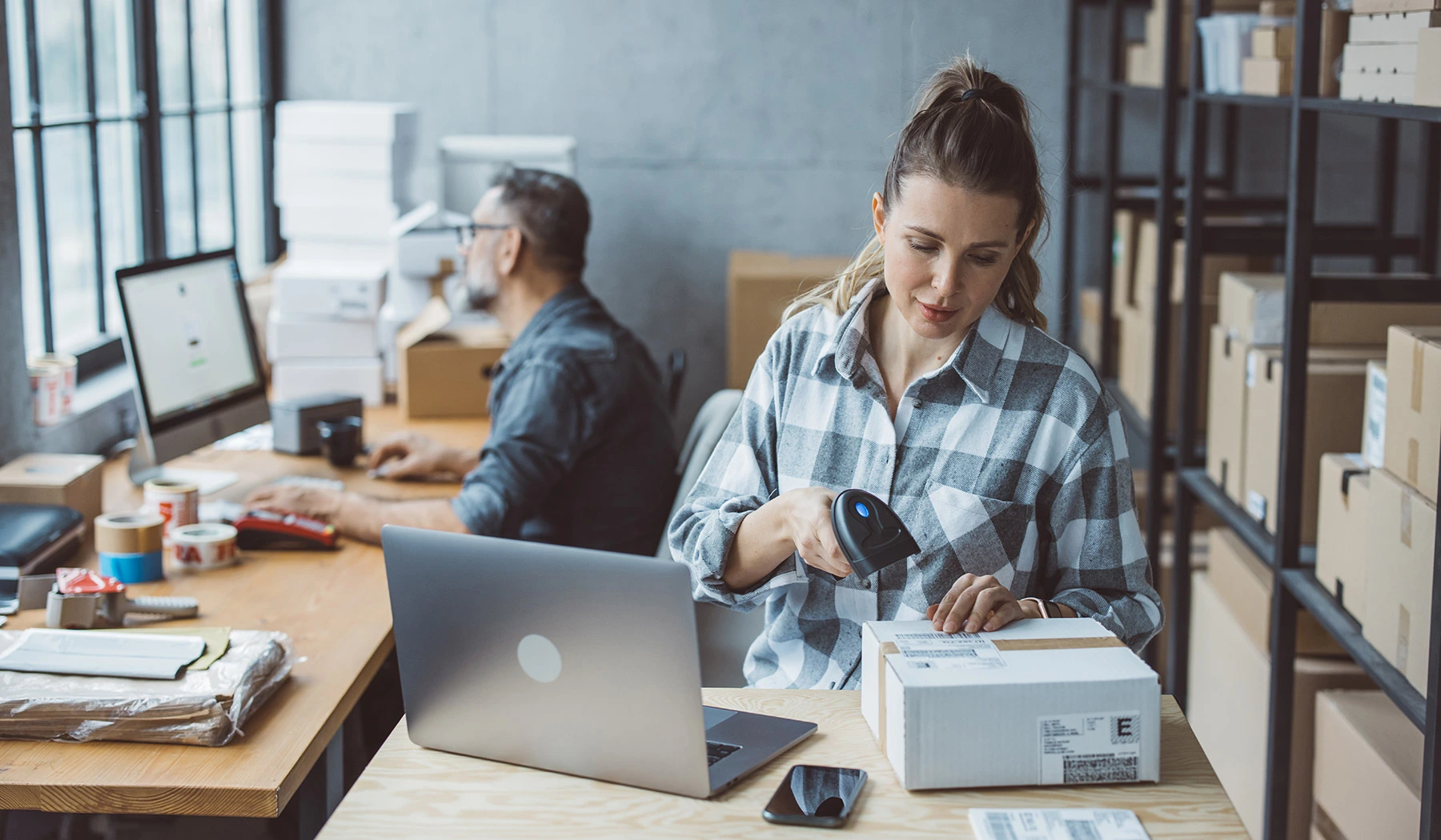 A worker in a checked shirt scans a shipping label on a cardboard box with a handheld barcode scanner at a fulfillment desk, while a colleague works at a computer behind them and shelves of packed boxes line the wall.